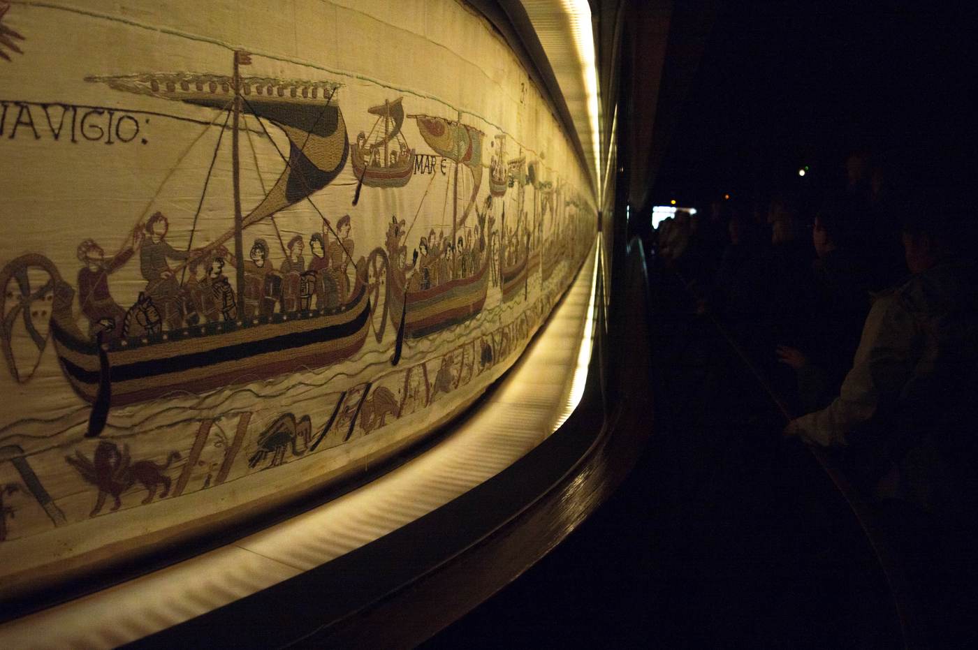 The Bayeux Tapestry displayed in its long illuminated case, with a visitor studying the embroidered ships and figures